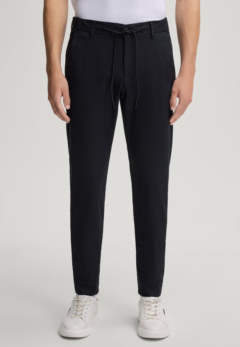 Granatowe spodnie z elastycznym paskiem w talii, sznurkiem do ściągania i mankietami przy kostkach. Gładki materiał z bocznymi kieszeniami. Noszone z białymi trampkami.