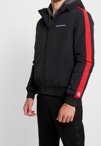 Svart vadderad jacka med huva, röda sidoremsor och "Calvin Klein Jeans"-logotyp. Har ribbade manschetter och elastisk nederkant.