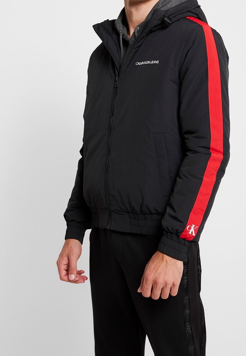 Svart vadderad jacka med huva, röda sidoremsor och "Calvin Klein Jeans"-logotyp. Har ribbade manschetter och elastisk nederkant.