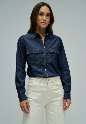 Mujer con camisa oscura azul con botones y bolsillos en el pecho y pantalones de talle alto color blanco roto, de pie frente a un fondo liso.