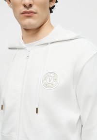 Νεαρό άτομο που φοράει λευκή φούτερ με φερμουάρ, με το λογότυπο Versace Jeans Couture και κορδόνια με χρυσές άκρες.