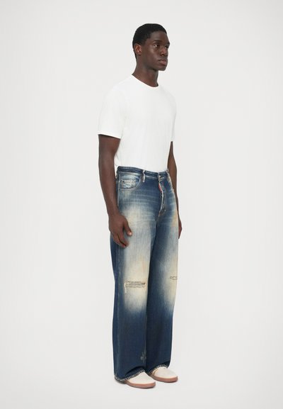 Uomo che indossa una t-shirt bianca, jeans larghi blu sbiaditi con strappi alle ginocchia e scarpe bianche, in piedi di fronte a uno sfondo chiaro e uniforme.