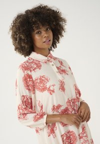 Bluză florală în crem cu modele de trandafiri roșii, având un guler cu nasturi și mâneci lungi cu manșete elastice.