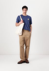 Blue short-sleeve t-shirt with sun graphic, beige pants, and brown shoes. Beige tote bag held in left hand, plain backdrop.