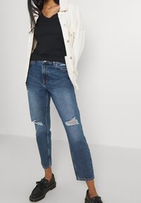 Mörkblå denimjeans med slitna hål, kombinerat med en svart V-ringad topp och en ljus oversized jacka. Svarta loafers fulländar looken.