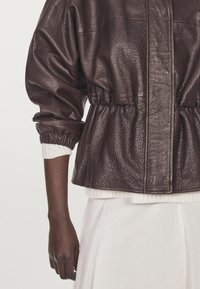 Burgunderfärgad läderjacka med samlad midja, voluminösa ärmar, mjuk textur och en enkel framknäppning, över en vit topp.