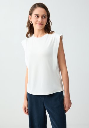 Femme aux cheveux bruns portant un haut blanc sans manches et un pantalon bleu foncé, regardant légèrement sur le côté avec un fond neutre.