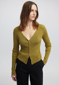 Cardigan a coste verde oliva con scollo a V, maniche lunghe e bottoni decorativi sul davanti, caratterizzato da una silhouette aderente e un orlo inclinato.