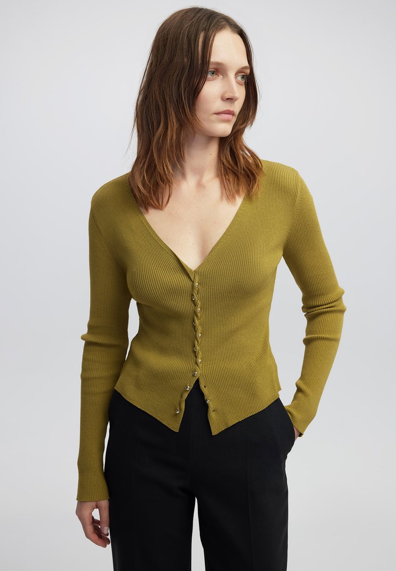Cardigan a coste verde oliva con scollo a V, maniche lunghe e bottoni decorativi sul davanti, caratterizzato da una silhouette aderente e un orlo inclinato.