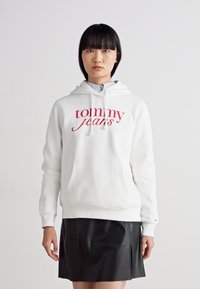Vit luvtröja med röd "tommy jeans"-logotyp, framficka och ribbade manschetter, tillsammans med en svart skinnkjol.