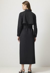 Zwarte lange jurk met een korte jasje overlay, getailleerde taille, lange mouwen en details met zijsplitten. Gladde textuur en minimalistisch ontwerp.