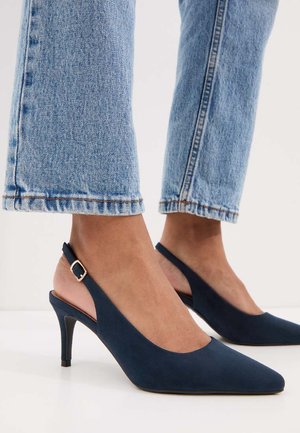 Voeten met marineblauwe slingback kittenheels gecombineerd met lichtblauwe spijkerbroeken tegen een witte achtergrond.