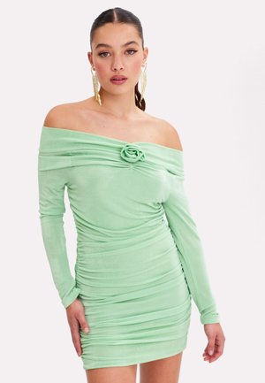 Vrouw in een muntgroene off-shoulder jurk met plooien, een bloemdetail bij de halslijn en lange mouwen, poserend tegen een lichte achtergrond.