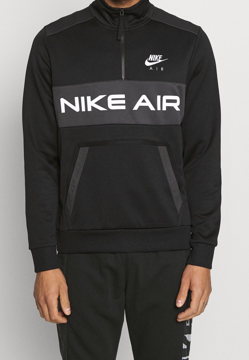 Homme portant un sweats à capuche noir Nike Air à demi-zip avec poche kangourou à l'avant et un pantalon noir Nike assorti.