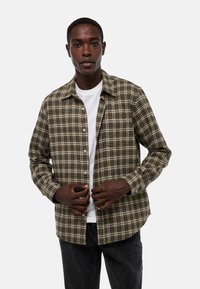 Chemise à carreaux marron et noir avec un col, une fermeture à boutons, des manches longues et une poche poitrine, portée sur un t-shirt blanc.