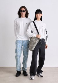 Două modele stau côte à côte. Primul poartă o cămașă albă cu mânecă lungă, având un logo verde, o pereche de pantaloni cu un model în nuanțe de albastru deschis și pantofi negri. Al doilea model este îmbrăcat cu o cămașă albă cu mânecă lungă, blugi negri largi, o geantă de umăr cu un model alb-negru și adidași negri.
