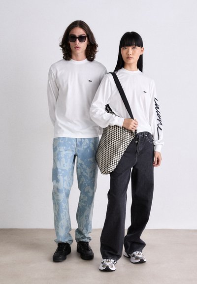 Deux modèles se tiennent côte à côte. Le premier porte une chemise blanche à manches longues avec un logo vert, un pantalon à motifs bleu clair et des chaussures noires. Le deuxième modèle a une chemise blanche à manches longues, un jean noir ample, un sac à bandoulière à motifs noir et blanc, et des baskets noires.