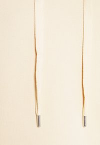 Gros plan sur un tissu de couleur crème montrant deux cordons suspendus avec embouts métalliques.