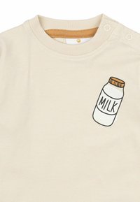 Helle beige Kurzarmbluse mit Rundhalsausschnitt und zwei Druckknöpfen, versehen mit einem bedruckten Milchflaschenmotiv auf der linken Seite.