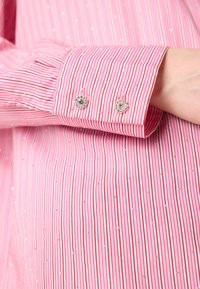 Striped shirt in pink with silver accents. Features vertical red and white stripes and decorative jeweled buttons on cuffs. Smooth texture.