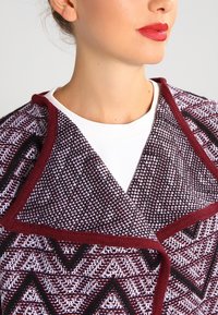 Stickad vinröd och vit sjal med ett geometriskt mönster, bred krage och texturerad tyg, buren över en vit topp.