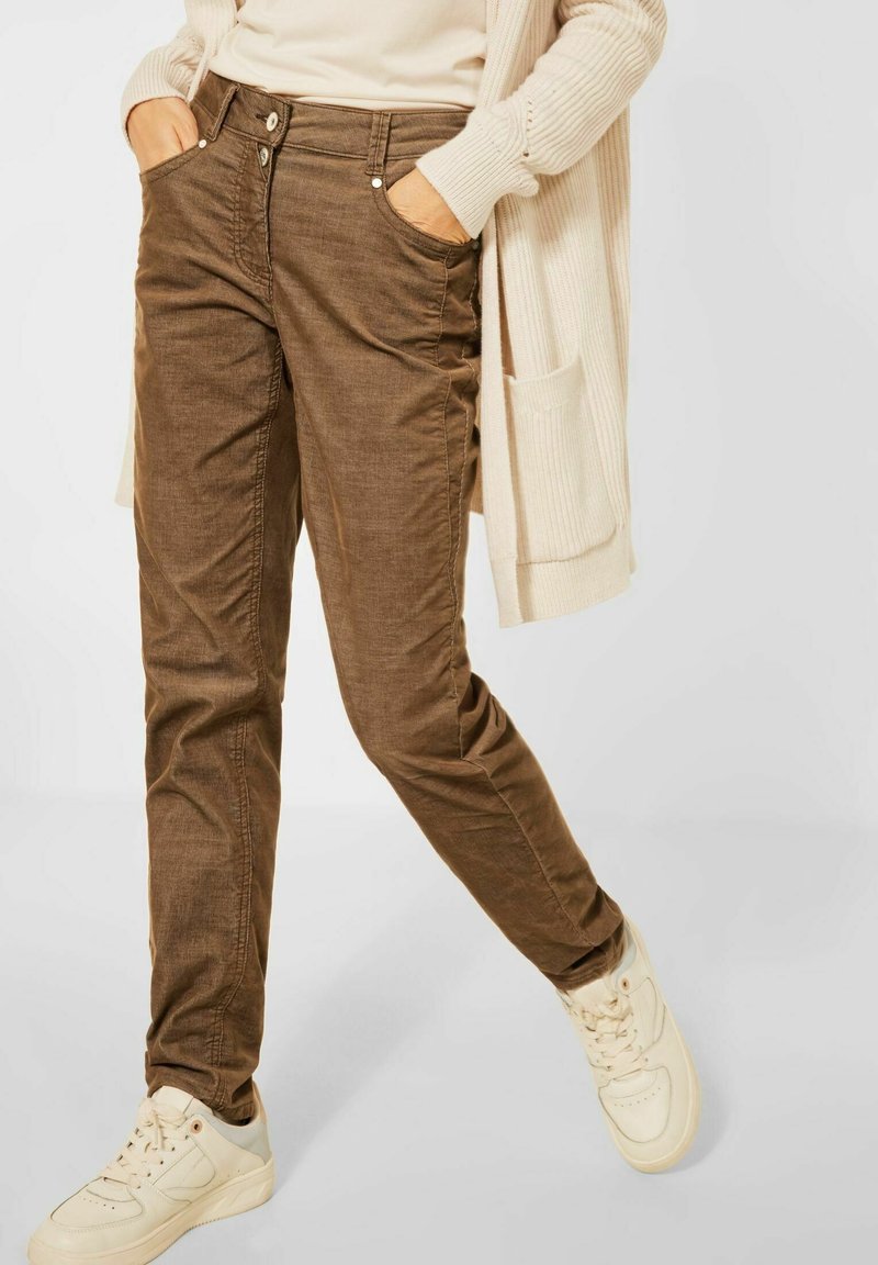 Bruna bomullsmixbyxor med slim fit, framfickor och knappstängning, kombinerat med ljusa sneakers mot en neutral bakgrund.