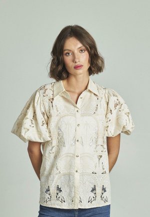 Blusa in pizzo color crema con chiusura a bottoni frontale, maniche corte a sbuffo e intricati motivi floreali. Presenta un colletto classico e un tessuto testurizzato.