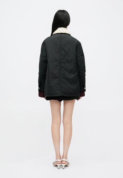 Sort oversized jakke med krave af hvidt shearling, præget af mørke manchetter og en glat stoffunktion. Vist fra ryggen, med shorts og sandaler.