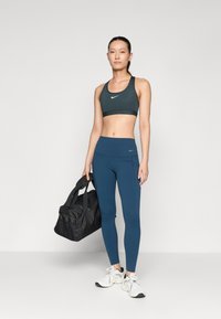 Tumši zaļš sporta krūšturis un zili legingi, abi ar Nike logo. Modelis tur melnu sporta maisu un valkā baltas sporta kurpes.