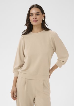 Femme portant un haut beige à manches longues bouffantes et un pantalon taille haute beige assorti, debout devant un fond clair uni.