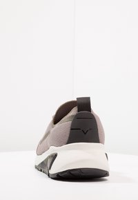 Lekki sneaker z teksturowaną, szarobrązową siateczką na cholewce, czarnym gumowym akcentem na pięcie i miękką białą podeszwą. Z tyłu znajduje się pętla do łatwego zakładania.