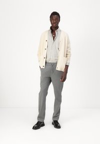 Cárdigan de punto crema con botones grandes, camisa a cuadros gris, pantalones grises y zapatos negros. El modelo está de pie contra un fondo blanco.