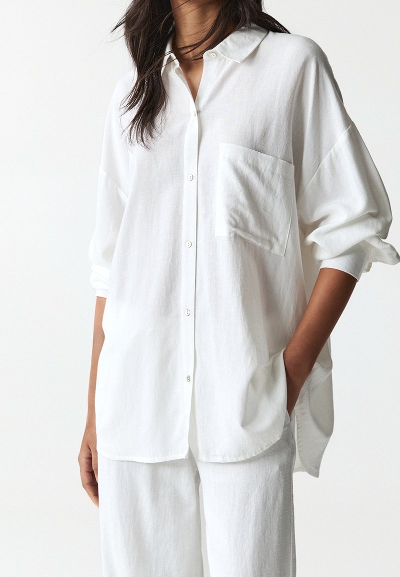 Femme portant une chemise blanche ample à manches longues boutonnée avec une poche et un pantalon blanc assorti, une main dans la poche.