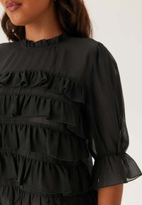 Blusa nera con design a strati e volant, tessuto trasparente, colletto alto e maniche a sbuffo con polsini elasticizzati.