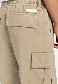 Pantaloni cargo beige con vita elasticizzata, tasche laterali e due tasche posteriori con pattina. Il materiale è resistente, con dettagli di cucitura visibili.