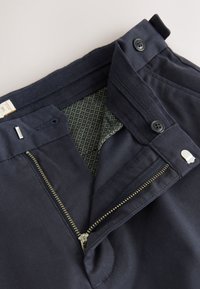 Pantalon bleu marine avec une fermeture éclair en métal, deux boutons noirs et une doublure intérieure verte texturée avec un motif géométrique.