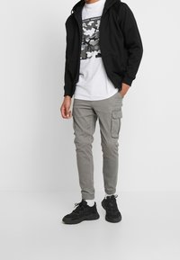 Musta vetoketjullinen huppari valkoisen T-paidan päällä, jossa on harmaa maastokuvio, yhdistettynä harmaisiin cargo-housuihin ja mustiin tennareihin.