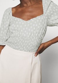 Blusa verde claro con lunares, mangas abullonadas y escote cuadrado; cuenta con una cintura fruncida. Combinada con una falda beige clara.