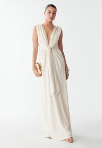 Dolga, brezrokavna krema obleka z globokim V-izrezom in vozlovnim detajlom na pasu. Ima teksturiran vzorec in subtilno drapiranje. Dopolnjuje jo zlata torbica.