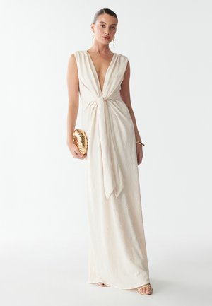 Dlouhé bezramínkové krémové šaty s hlubokým výstřihem ve tvaru V a detailem uzlu v pase. Mají texturovaný vzor a jemné drapování. Doplněny zlatou kabelkou.
