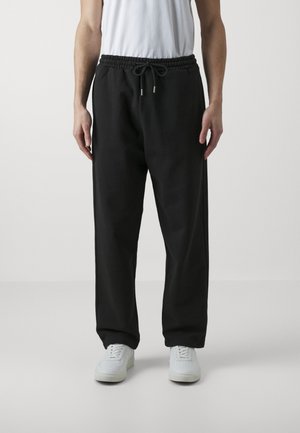 Fekete melegítőnadrág húzózsinórral a derekán, szűkülő lábszárral és oldalsó zsebekkel, fehér sportcipővel egy egyszínű háttér előtt.