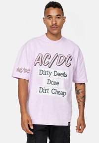 Světle fialové bavlněné tričko s černým a růžovým textem: "AC/DC Dirty Deeds Done Dirt Cheap." Volný, oversized střih s krátkými rukávy.