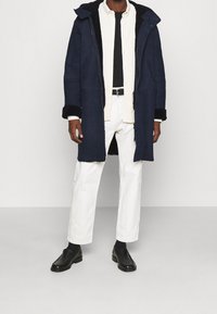 Parka bleu marine avec capuche bordée de fourrure, devant boutonné, et poignets noirs, portée sur une chemise blanche et une chemise noire, associée à un pantalon blanc.