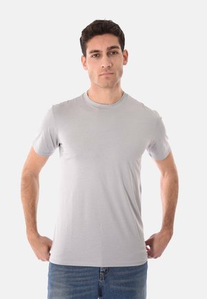 Giovane uomo con capelli ricci scuri che indossa una maglietta a maniche corte grigio chiaro e jeans blu, in piedi con le mani vicino ai fianchi.