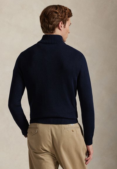 Mężczyzna o krótkich brązowych włosach nosi granatowy sweter z golfem i beżowe spodnie, stoi tyłem na neutralnym tle.