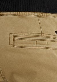 Chinos kaki avec une texture lisse, dotés d'une poche arrière avec des détails cousus. Ceinture élastique noire visible en haut.
