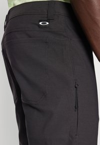Pantalons noirs en tissu léger, dotés d'une poche arrière, d'un cordon de serrage ajustable et d'un patch logo distinctif. Texture lisse, design minimaliste.