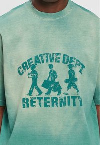Grøn oversized t-shirt lavet af bomuld. Den har et slidtegn på trykket af tre figurer med teksten "CREATIVE DEPT. RETERNITY" nedenunder.