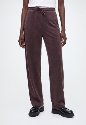 Personne portant un pantalon en velours côtelé marron foncé, ample et droit, avec une taille à cordon de serrage et des chaussures noires épaisses sur un fond clair uni.