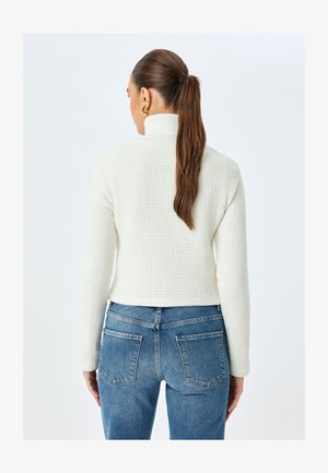 Weißer, strukturierter Pullover mit hohem Kragen, kombiniert mit hellblauen Jeans. Der Pullover hat lange Ärmel und einen cropped Saum.